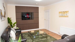 Een woonkamer met een leren bank, een glazen salontafel, een televisie bevestigd aan een houten wandpaneel en een deur aan de rechterkant.
