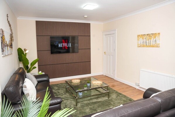 Un salón con sofá de cuero, una mesa de centro de vidrio, un televisor montado en un panel de pared de madera y una puerta a la derecha.