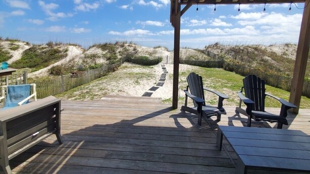 Een houten terras met Adirondack-stoelen en een pooltafel, met uitzicht op een zandstrand en duinen.
