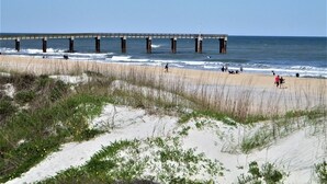 Een strand met een steiger die in het water uitloopt, zandduinen en mensen die langs de kust wandelen.