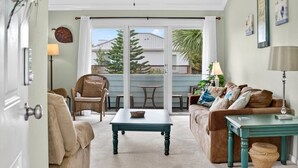 A living room with a sofa, a coffee table, and a dining table. There is a sliding glass door leading to a balcony with a view of trees and a fence. - Quail Hollow A8-1u 2 Bedroom Condo (St. Augustine)