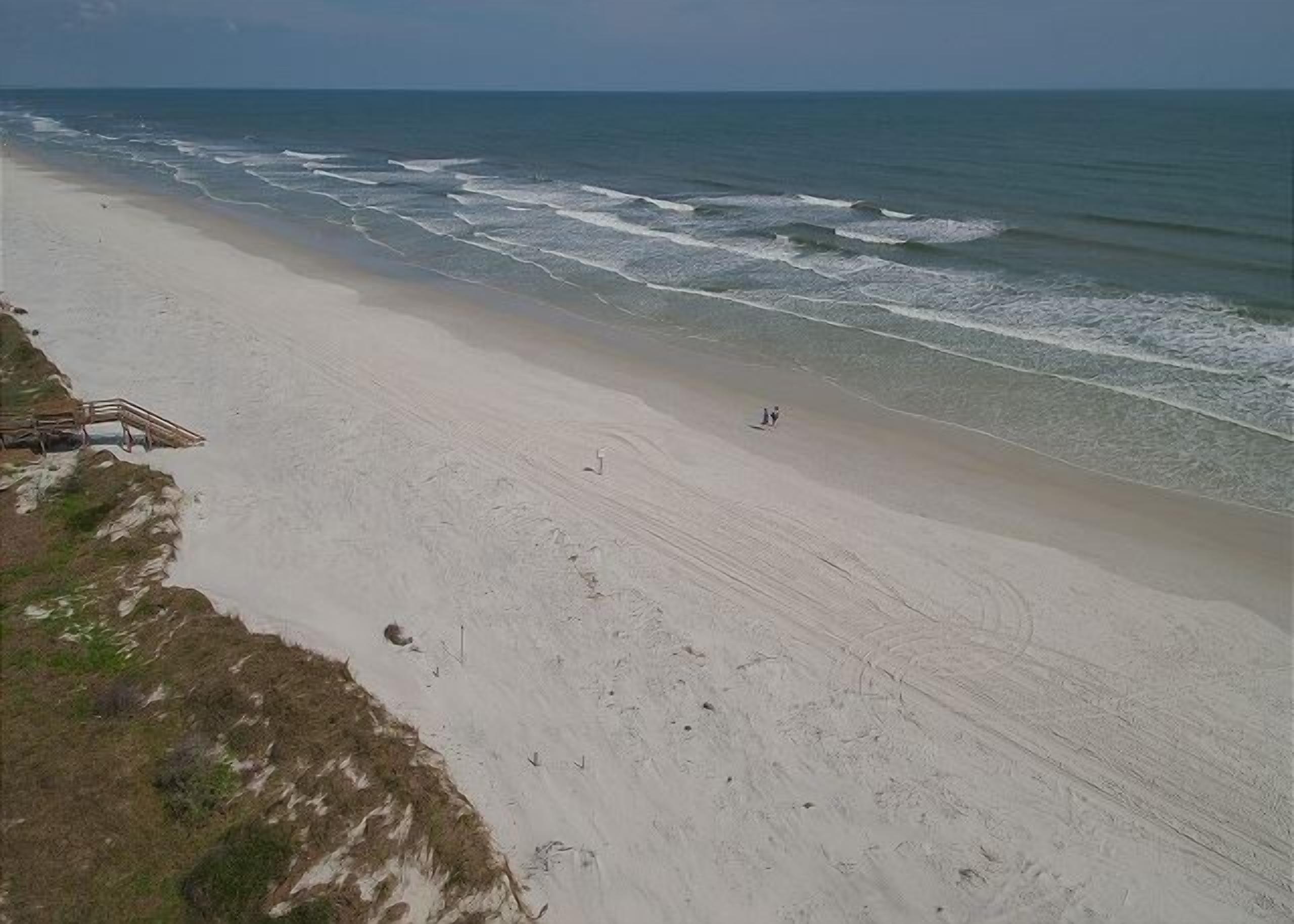 A beach with a wooden walkway leading to the water, a sandy shore, and a few people walking.