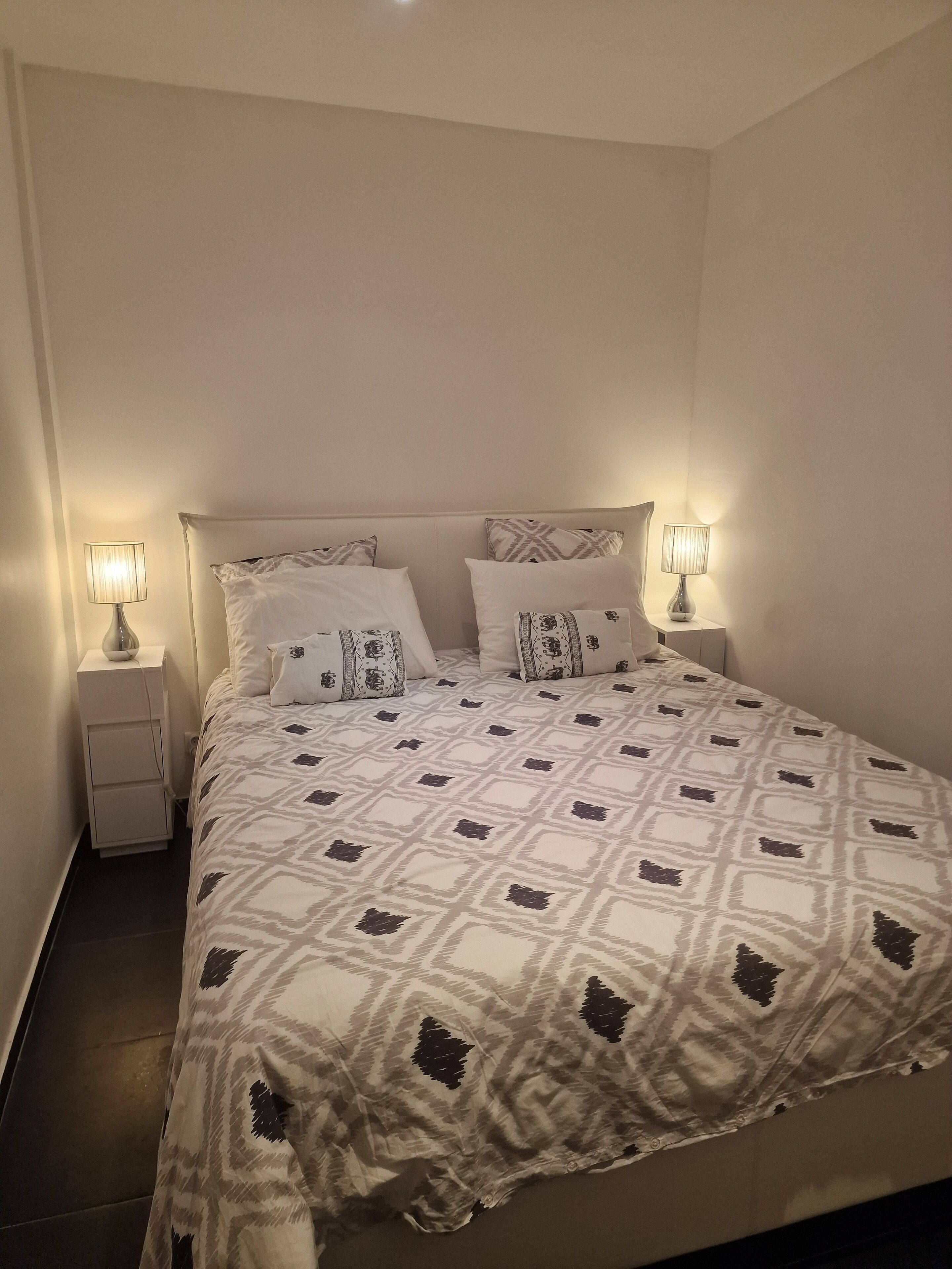 Un letto rifatto con cura, una coperta a motivi, due abat-jour e un comodino.
