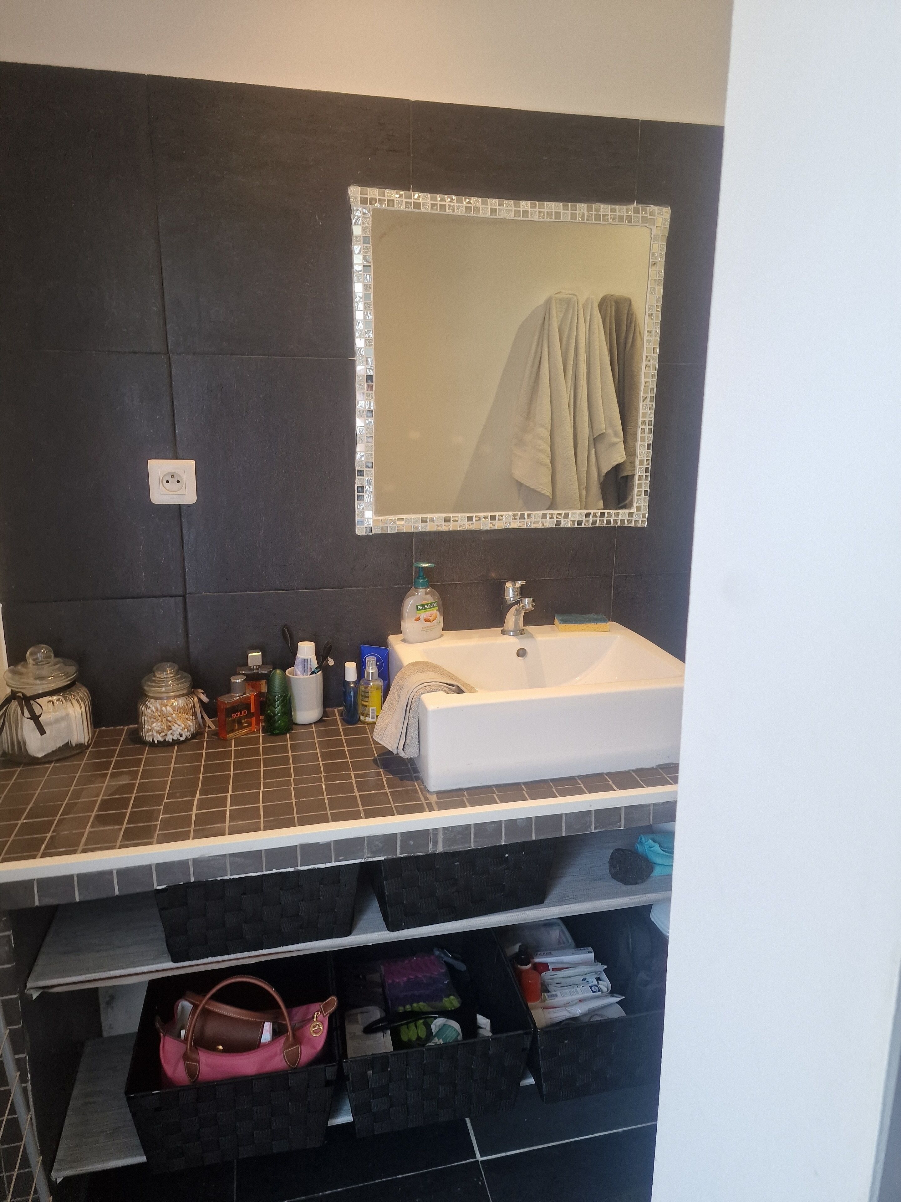 Une salle de bain avec un lavabo blanc, des murs recouverts de carreaux foncés, un miroir doté d’un cadre décoratif et une serviette accrochée au mur.