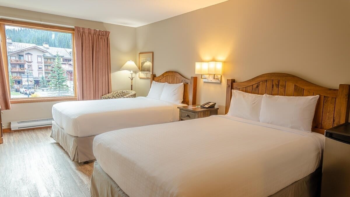 A hotel room with two beds, a window with a view of buildings and trees, a wooden headboard, bedside lamps, and a nightstand.
