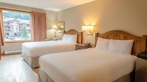 A hotel room with two beds, a window with a view of buildings and trees, a wooden headboard, bedside lamps, and a nightstand.