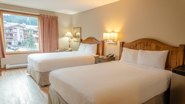 A hotel room with two beds, a window with a view of buildings and trees, a wooden headboard, bedside lamps, and a nightstand. - Cahilty #212 Hotel Room (Sun Peaks)