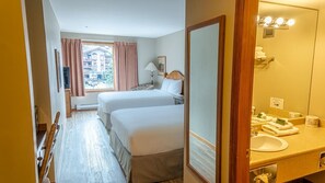 A hotel room with two beds, a large mirror, a bathroom with a sink and towel rack, and a window with curtains. - Cahilty #212 Hotel Room (Sun Peaks)