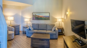 A modern living room with a grey sofa, two side tables with lamps, a wooden coffee table, a flat-screen TV, and a painting of a landscape on the wall.