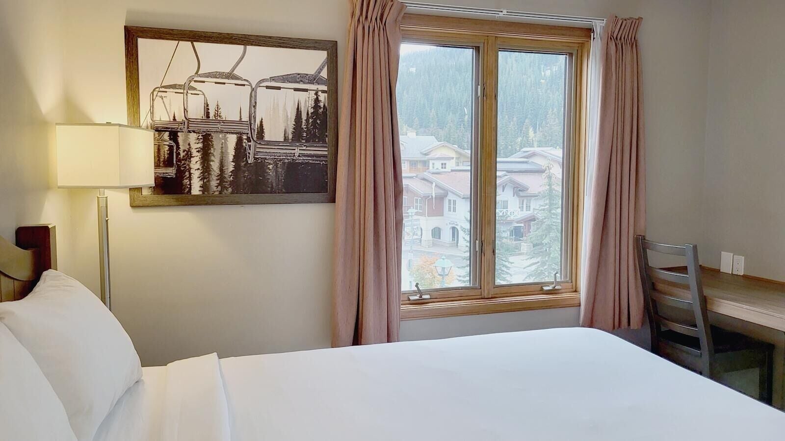 Una habitación de hotel con cama, escritorio, silla, lámpara y ventana con cortinas.