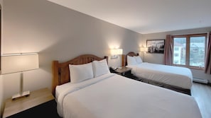 A hotel room with two beds, a nightstand, a lamp, and a window with curtains.