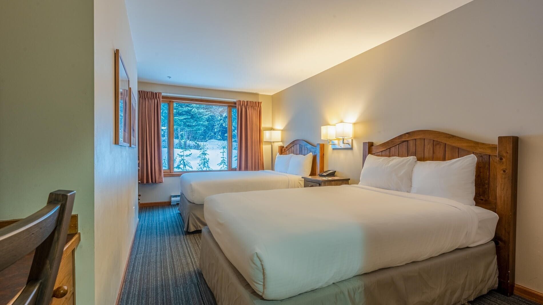 Habitación de hotel con dos camas, un ventanal grande con cortinas, un cabecero de madera y un pequeño escritorio con lámpara.