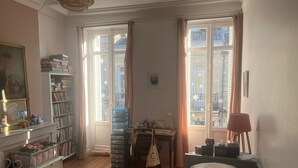 Et hyggeligt soveværelse med trægulv, en seng med hvidt sengetøj, et skrivebord med stol, en reol og et vindue med gardiner.