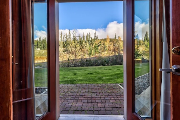 A wooden door leading to a paved area with a view of a forest and a grassy field.