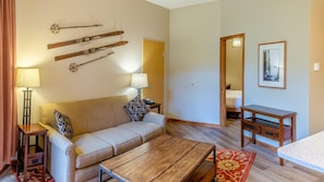 Uma sala de estar acolhedora com um sofá, mesa de centro e uma mesinha de cabeceira de madeira. Há esquis fixados na parede, um abajur e um quadro emoldurado.