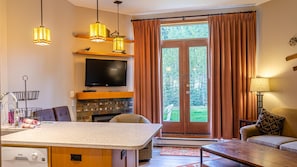 A modern living room with a flat-screen TV, a fireplace, a dining area with a kitchen island, and a large sliding glass door leading to a garden.