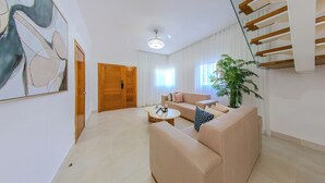 A spacious living room with a beige sofa, a coffee table, and a large plant. A wooden door and a staircase are visible in the background.