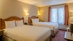 A hotel room with two beds, wooden headboards, bedside lamps, a window with curtains, and a carpeted floor.