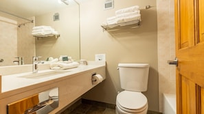 A modern bathroom with a sink, mirror, towel rack, toilet, and a wooden door. - Cahilty #261 Hotel Room (Sun Peaks)