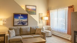 A living room with a sofa, armchair, and coffee table. There are framed pictures on the wall, a lamp, and a window with curtains.