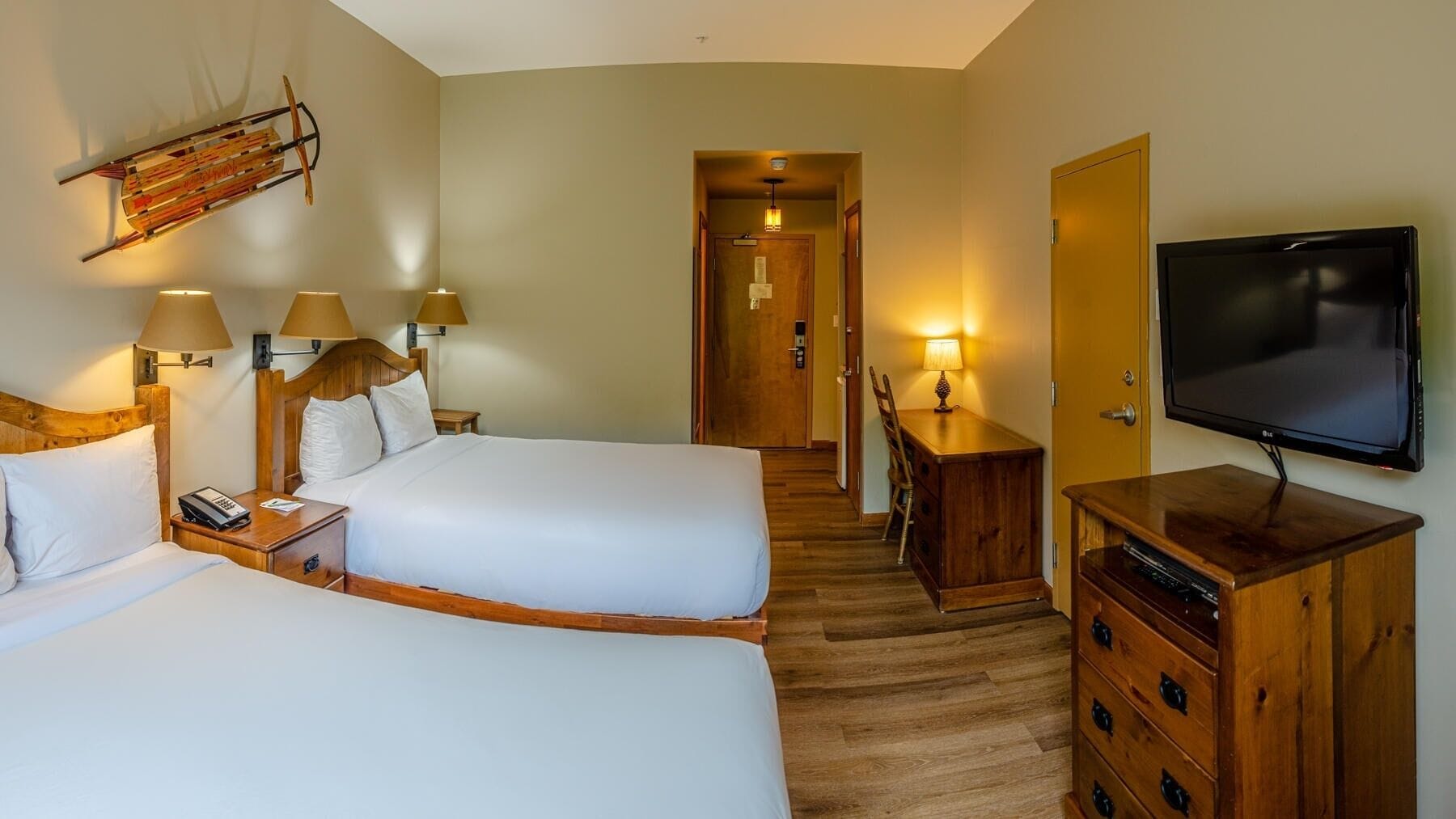 A hotel room with two beds, a TV, wooden furniture, and a hallway leading to a door.