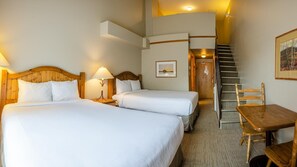 A hotel room with two beds, a wooden table, a chair, a staircase, and a framed picture on the wall.