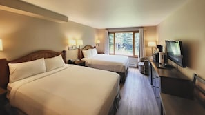 A hotel room with two beds, a TV, a desk, and a window with a view of trees.
