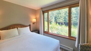 Una habitació d'hotel amb un llit gran, una tauleta de nit amb una làmpada i una finestra amb vista al bosc.