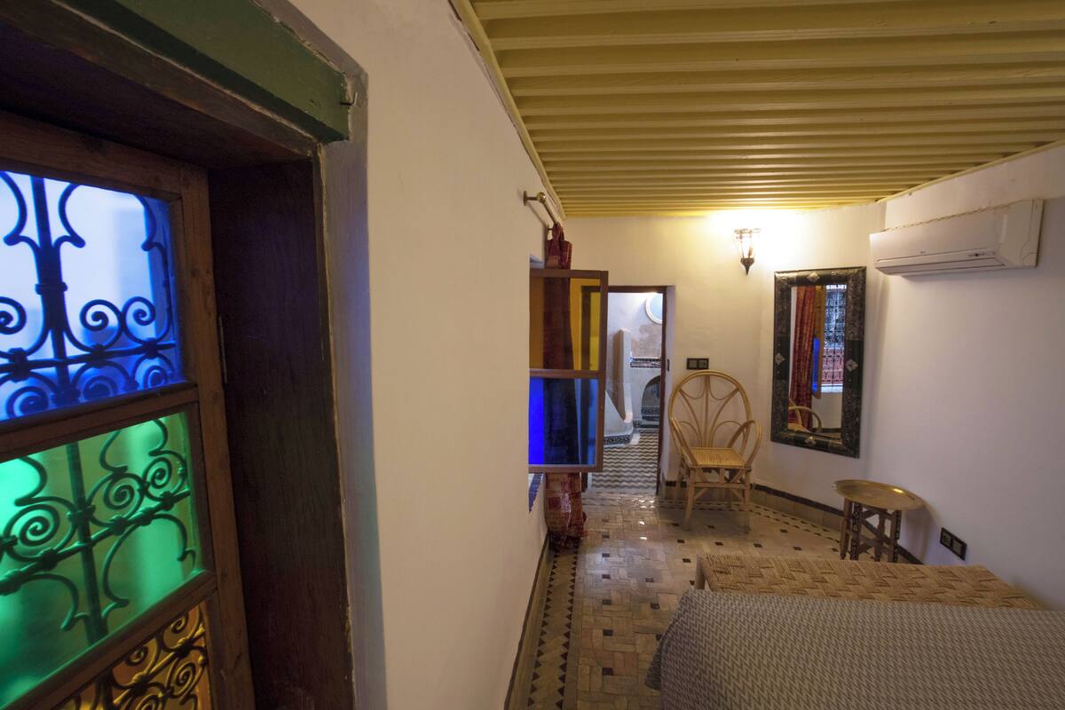 Un pasillo estrecho con suelo estampado, una puerta de madera con un vitral, una silla de mimbre, una pequeña mesa redonda y un equipo de aire acondicionado en la pared.
