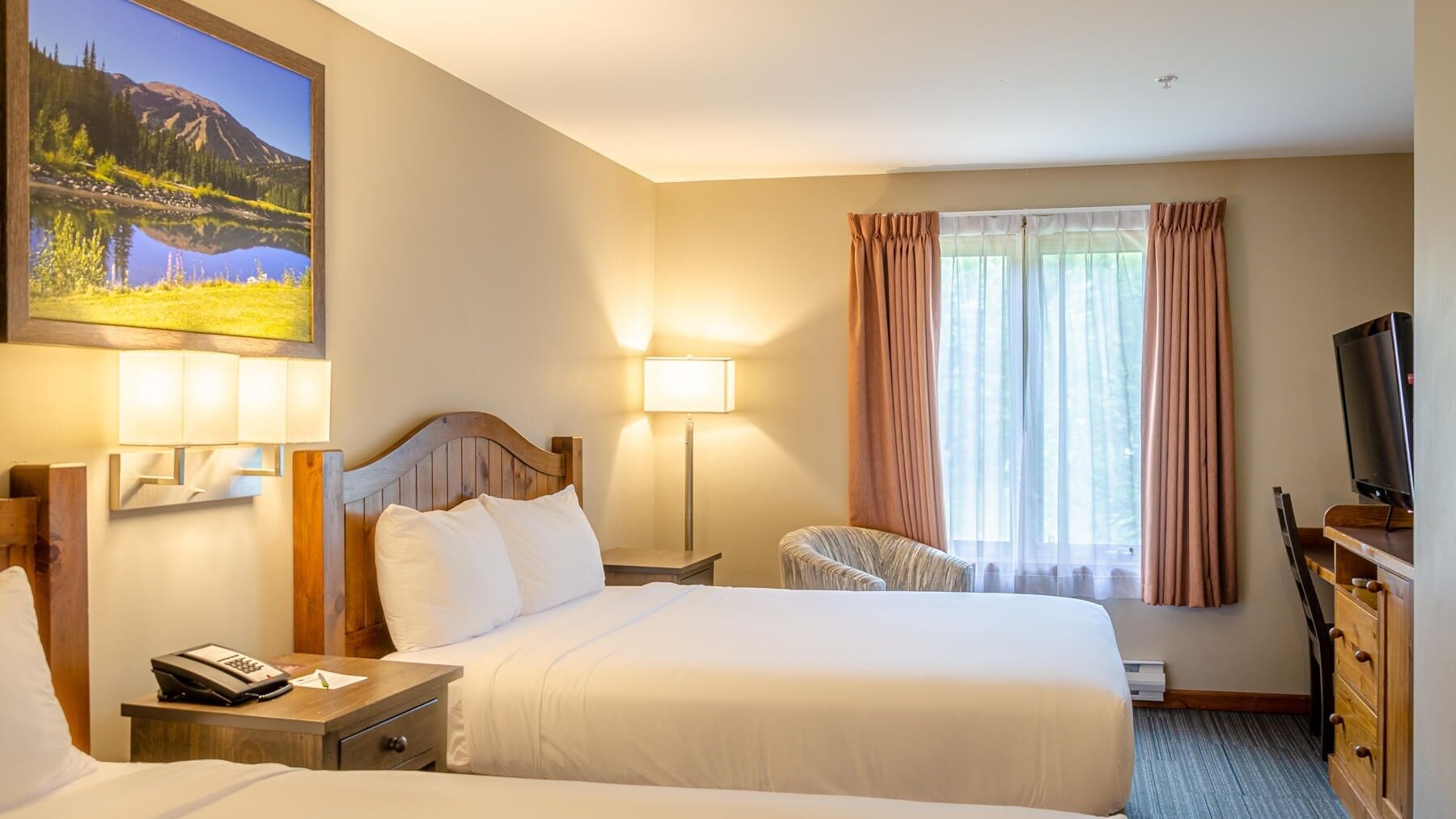 A hotel room with two beds, a desk with a lamp, a TV, and a painting of a mountain landscape.