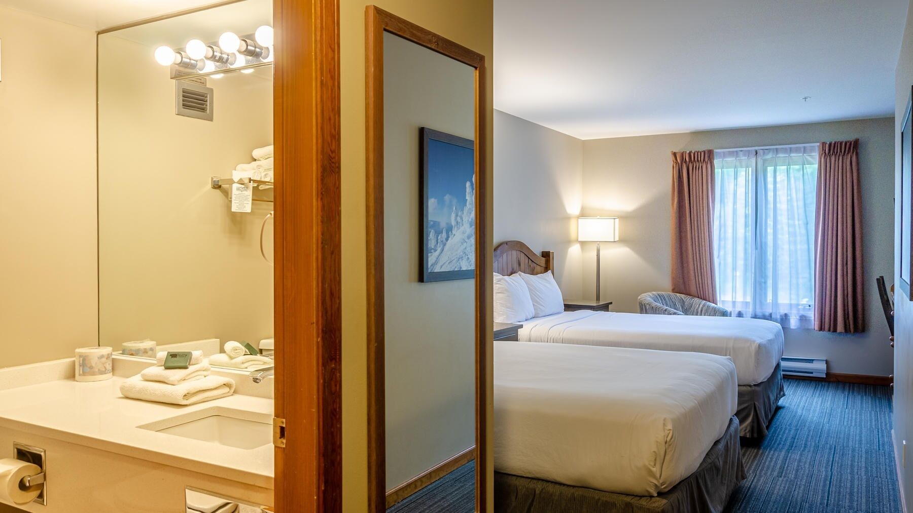 A hotel room with two beds, a bathroom with a mirror, and a window with curtains.