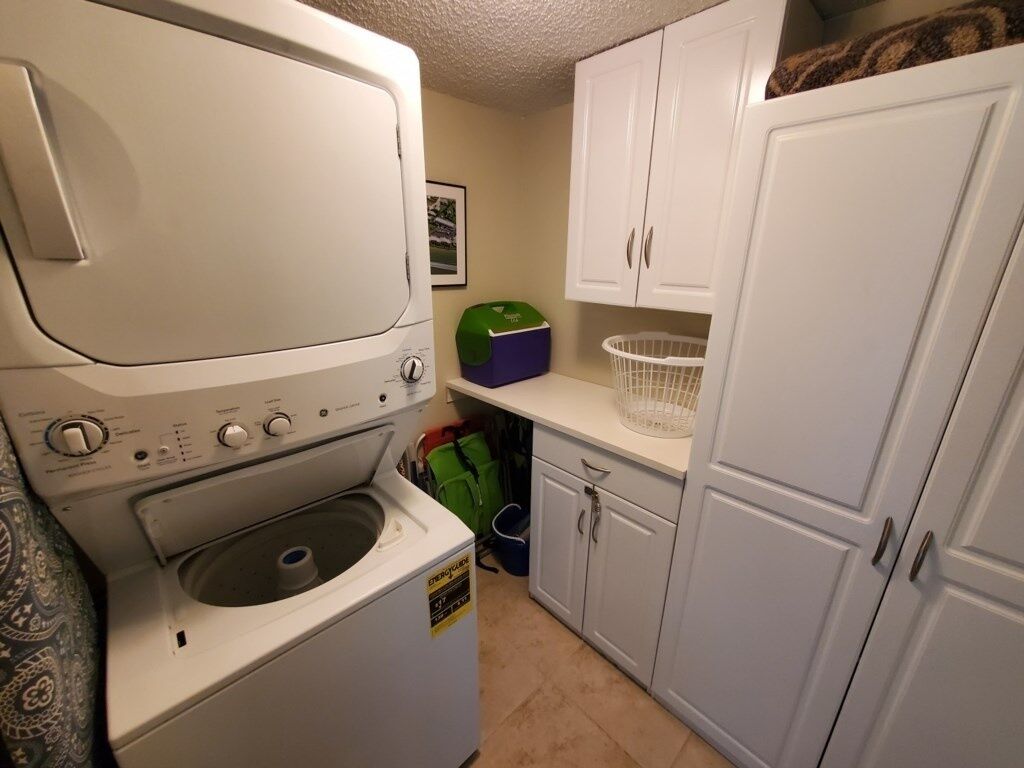 A compact laundry area with a front-loading washer and dryer, a green storage bin, and white cabinets.