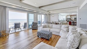 A spacious living room with a large window overlooking the sea, a cozy seating area, and a modern kitchen in the background. - Ocean Paradise 4 Bedroom Home (St. Augustine)
