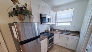 A compact kitchen with a stainless steel refrigerator, microwave, and oven, white cabinets, a granite countertop, and a window providing natural light. - Zen Cottage - All 4 Bedroom Home (St. Augustine)