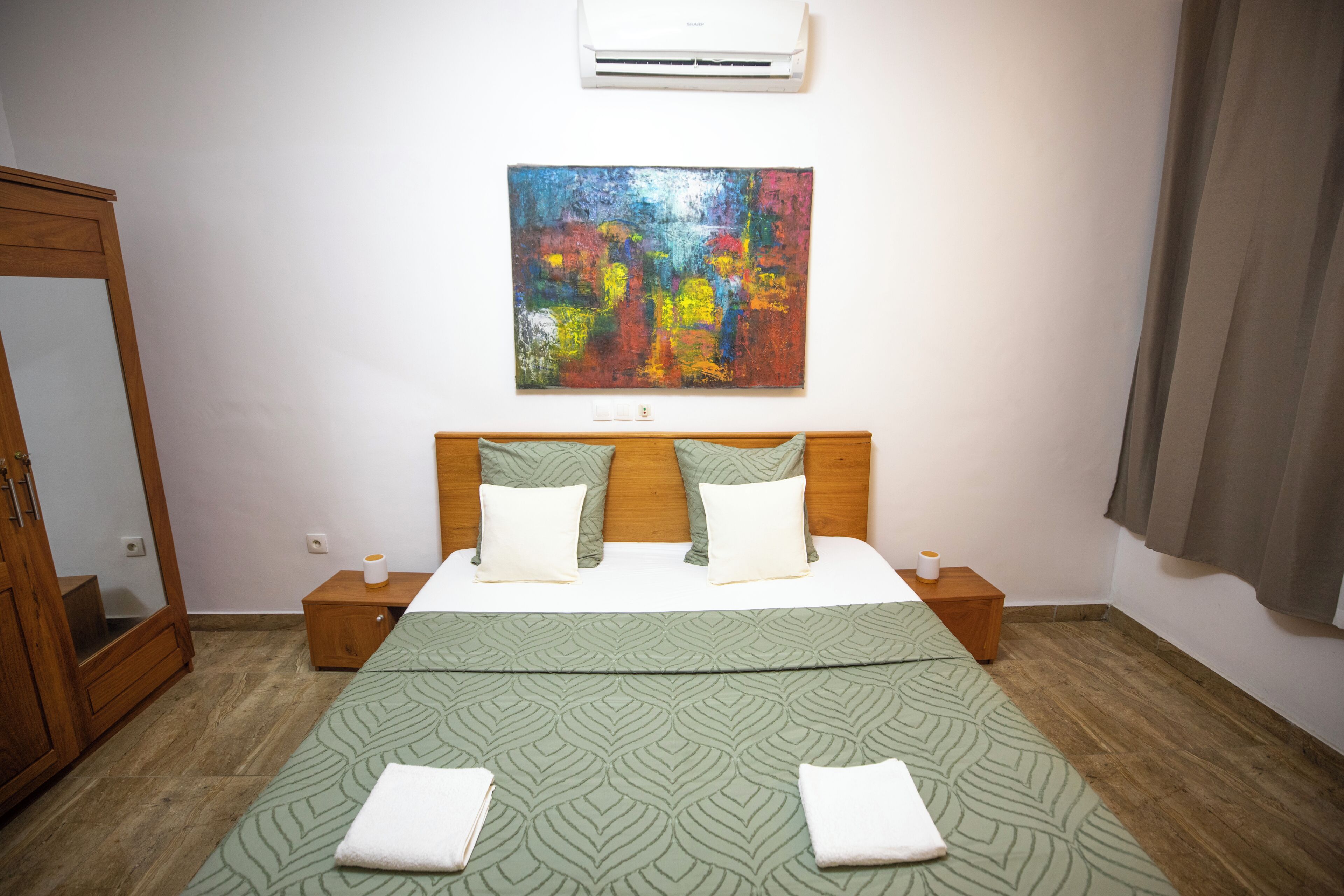 A bedroom with a bed, a nightstand, a mirror, an air conditioner, and a painting on the wall.