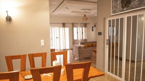 Un comedor con una mesa y sillas de madera, un techo con iluminación empotrada y vistas a una sala de estar con sofá y cortinas.