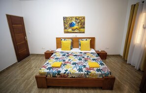 Une chambre à coucher avec un lit en bois, deux oreillers jaunes, une couvre-lit à motifs floraux, un tableau au mur et une porte en bois.