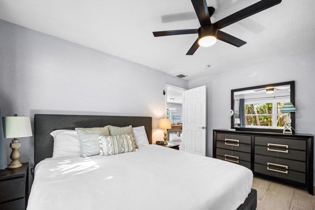 A bedroom with a bed, nightstands, a dresser, a ceiling fan, and a window with a view of greenery.