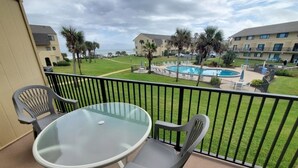 Un balcon doté d’une table en verre et de deux chaises, offrant une vue sur une piscine et un espace verdoyant agrémenté de palmiers, avec des bâtiments en arrière-plan.