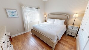 A bedroom with a large bed, a bedside table with a lamp, a picture on the wall, and a window with curtains. - Alerio 103e 2 Bedroom Condo (Miramar Beach)