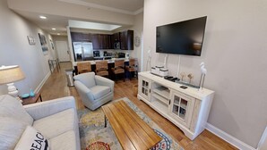 A living room with a sofa, armchair, and coffee table. There is a kitchen in the background with dark cabinets and a flat-screen TV mounted on the wall. - Alerio 103e 2 Bedroom Condo (Miramar Beach)