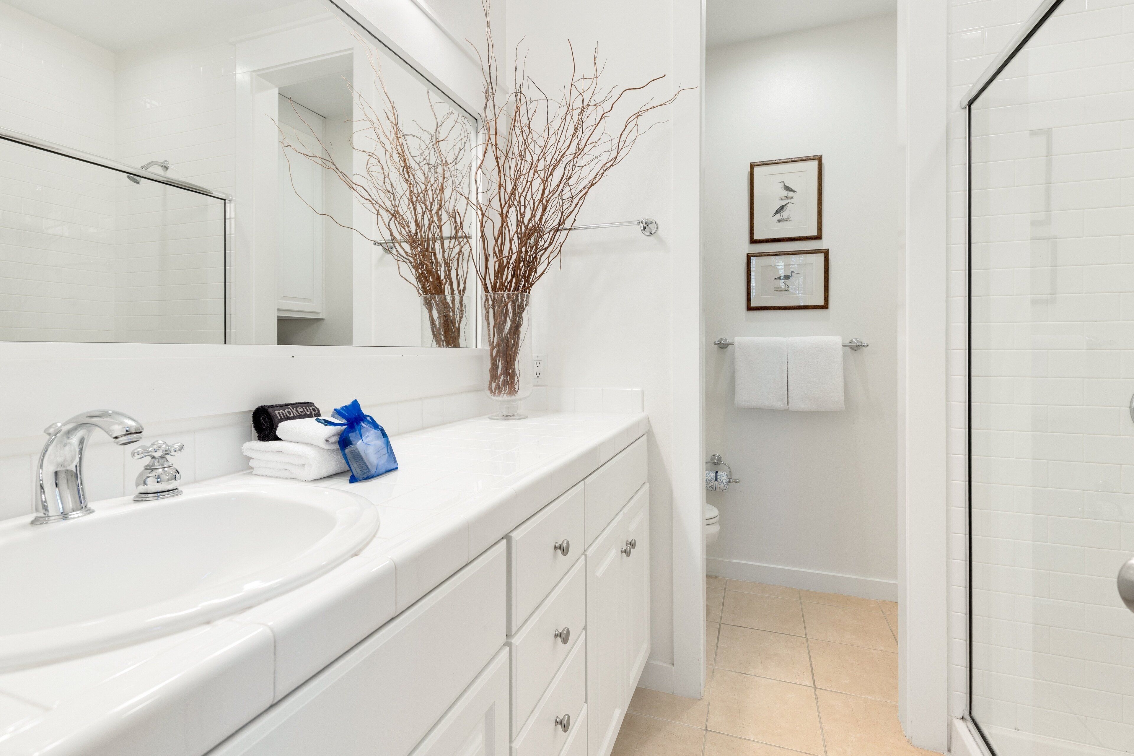 Un baño moderno con lavamanos, espejo y ducha blancos. Hay un florero con ramas en la encimera y dos cuadros en la pared.