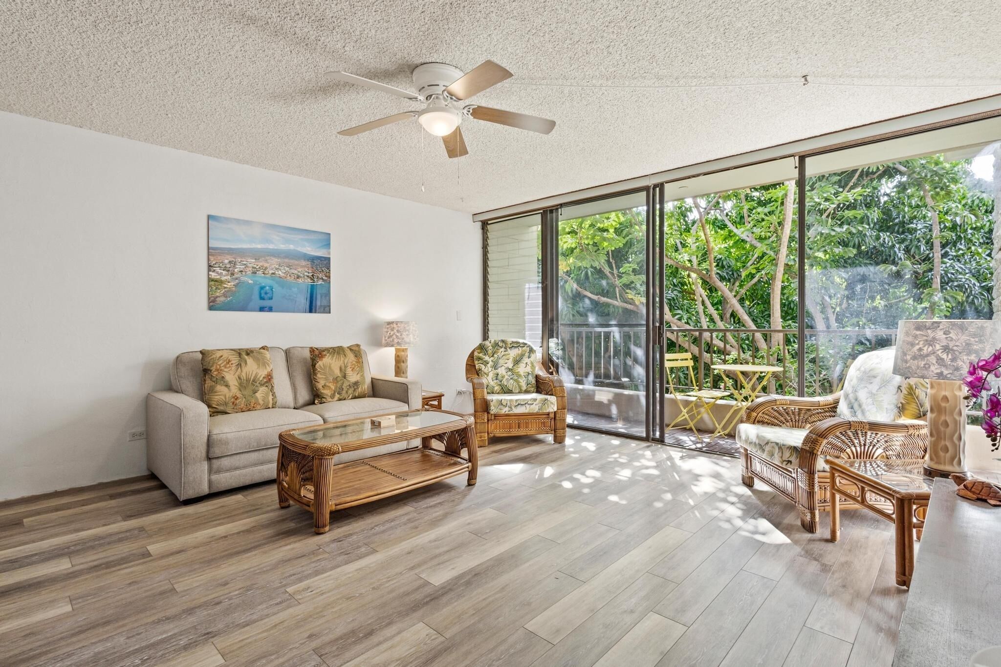 A spacious living room with a sofa, armchairs, a coffee table, and a ceiling fan. Large windows offer a view of greenery outside.