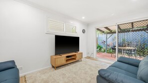 A living room with a flat-screen TV on a wooden stand, a blue sofa, and a sliding glass door leading to an outdoor patio with plants and furniture. - Flynns Beach Retreat, 3 of 3 Everard street (Port Macquarie)