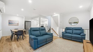 A modern living room with two blue sofas, a dining table with chairs, a kitchen in the background, and a staircase leading to another level. - Flynns Beach Retreat, 3 of 3 Everard street (Port Macquarie)