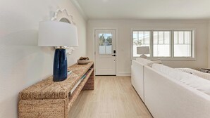 Et moderne, lyst rum med et stort vindue, en hvid sofa, en bænk med en lampe og en dør der fører ud.