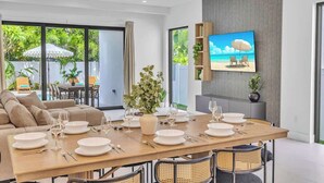 Un comedor con una mesa de madera preparada para seis personas, rodeada de sillas de mimbre, un sofá y un televisor montado en la pared.