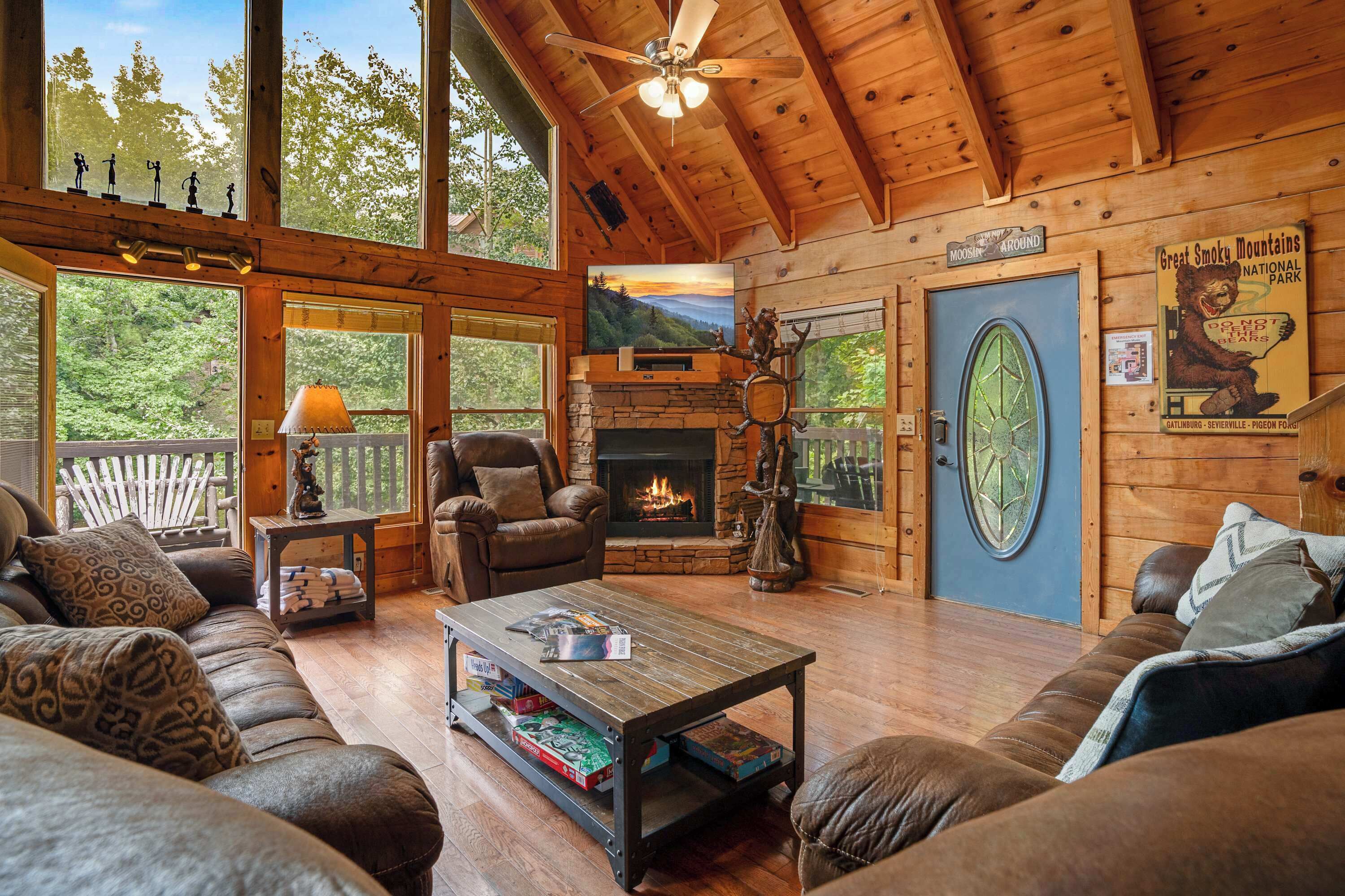 A cozy cabin interior with a fireplace, leather furniture, and a large window overlooking trees.