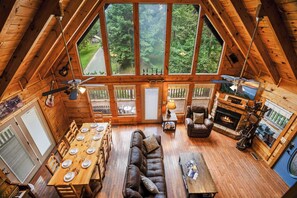 Un loft spacieux avec de hauts plafonds, de grandes fenêtres donnant sur une forêt, un coin repas avec une table et des chaises en bois, un canapé en cuir et une cheminée.