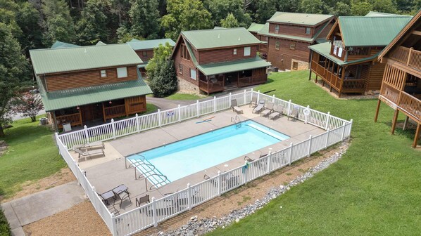 A pool area surrounded by wooden cabins with green roofs, a white fence, and outdoor seating. - Handicap Friendly Cabin Retreat・Pool Table・Grill (Pigeon Forge)